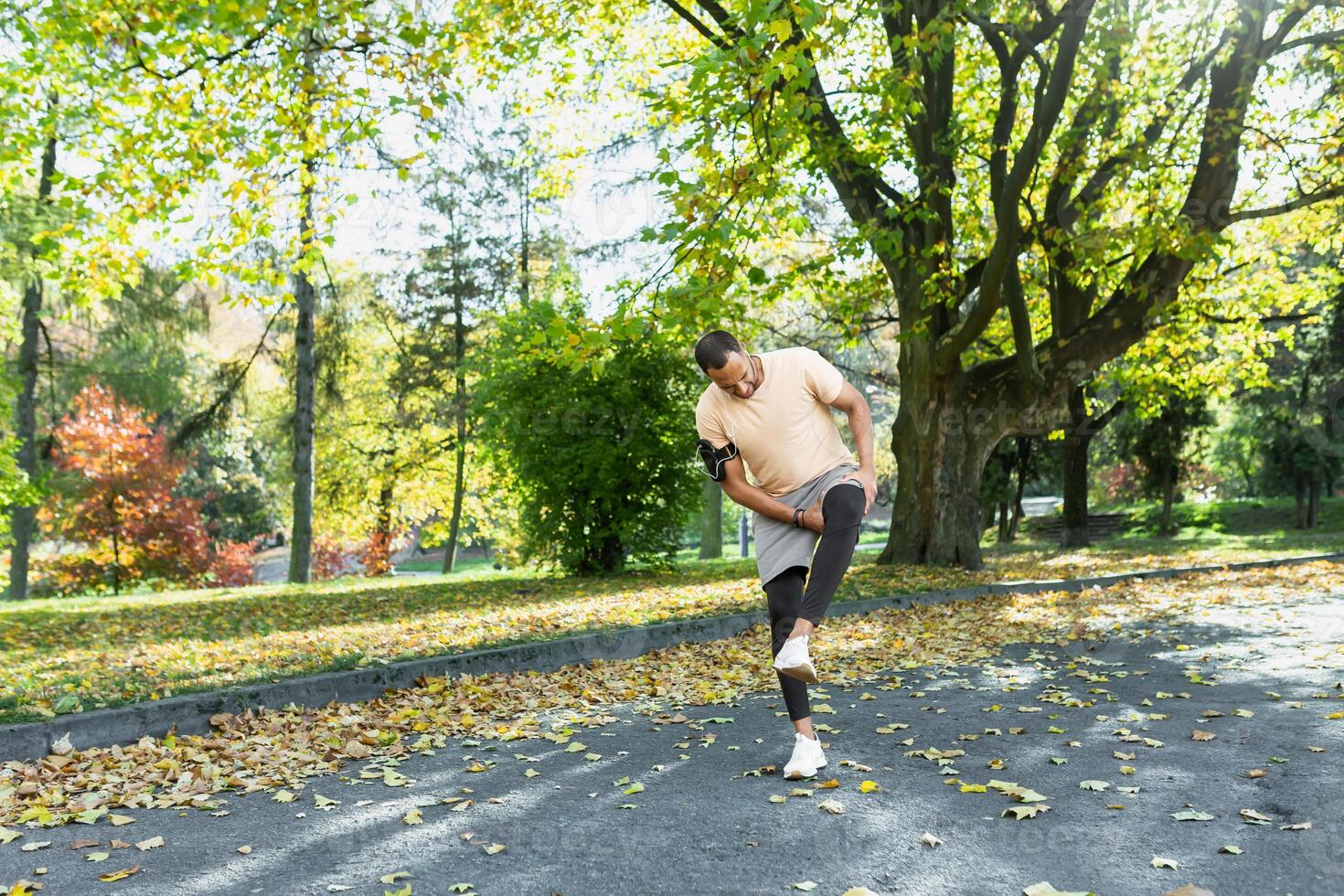 Runner injured his leg while jogging in the park near the trees, hispanic man has muscle spasm massaging his knee, man is running and exercising fitness on a sunny day. photo