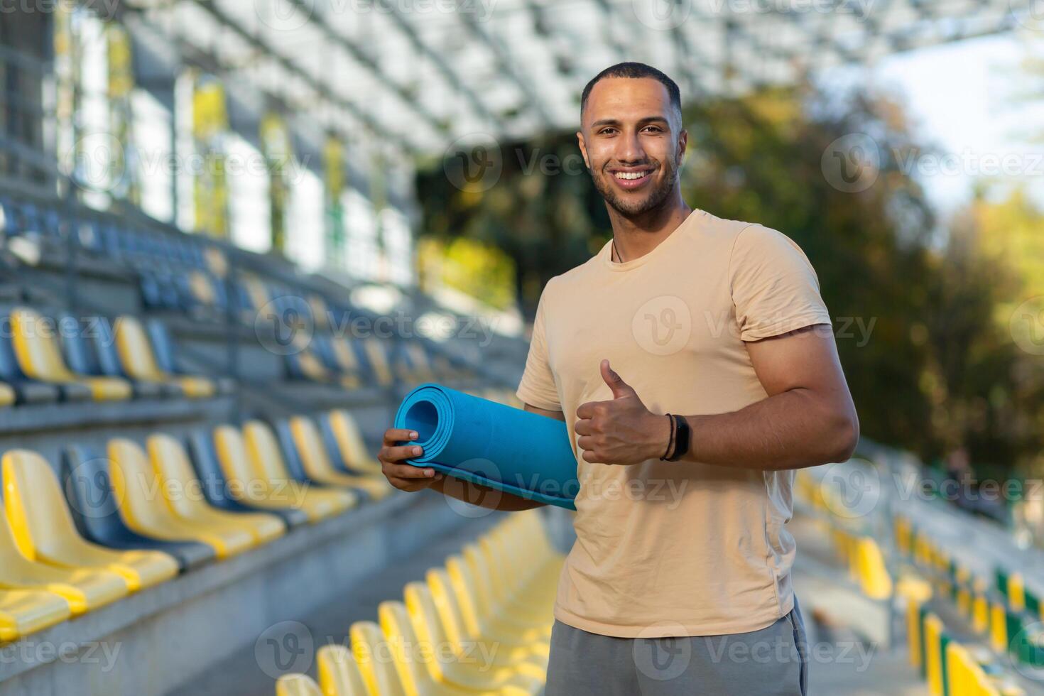 Portrait of hispanic male fitness trainer at sports stadium, athlete with yoga mat smiling and looking at camera, athlete in sportswear at sunset showing thumbs up. photo