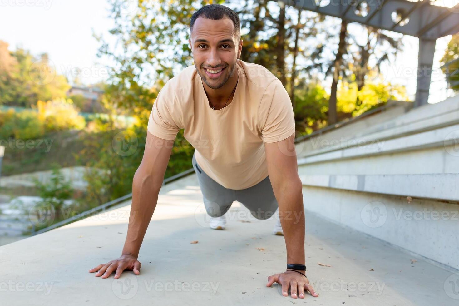 Cheerful and smiling hispanic man doing outdoor exercise in stadium, man stretching and recuperating before jogging in summer park. photo
