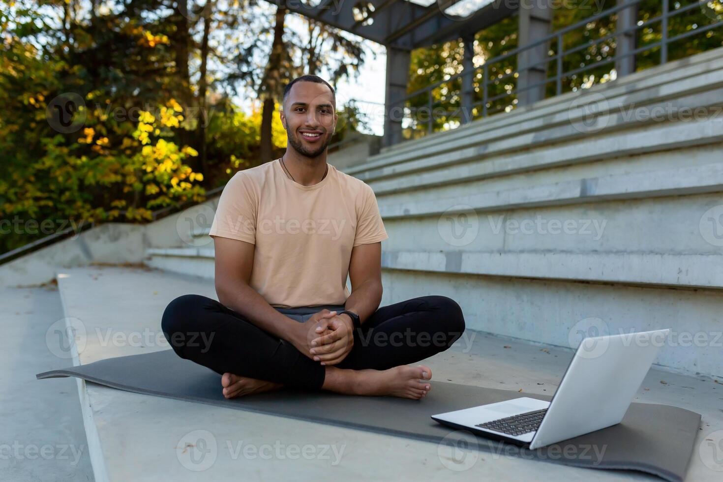 Portrait of arab sportsman fitness trainer, man smiling and looking at camera sitting on sports mat in stadium, using laptop to teach online course photo