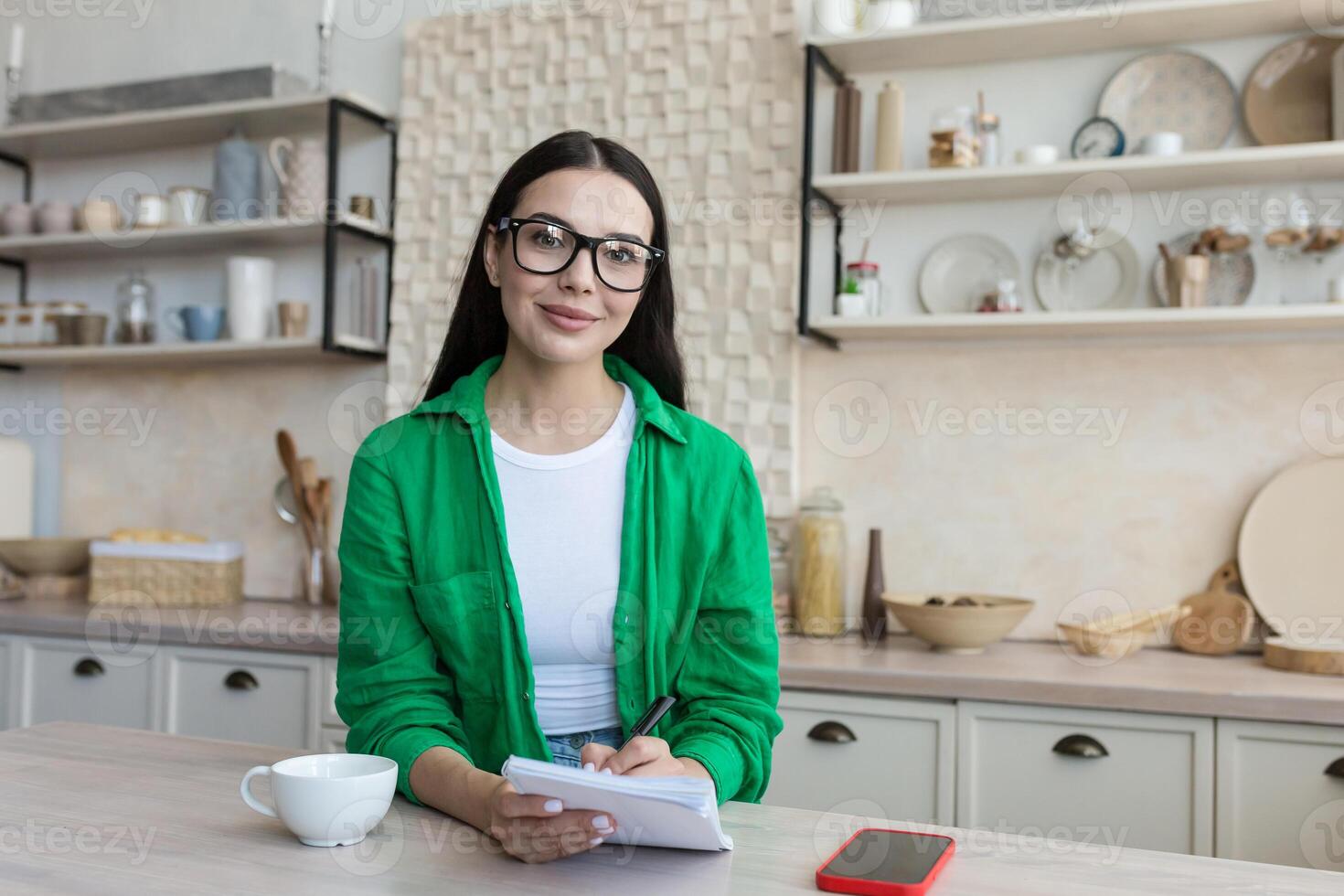 A young woman sits at home in the kitchen and writes with a pen in a notebook, keeps a personal diary, writes down dreams, makes a plan, works on self-developmente. He looks at the camera with a smile. photo