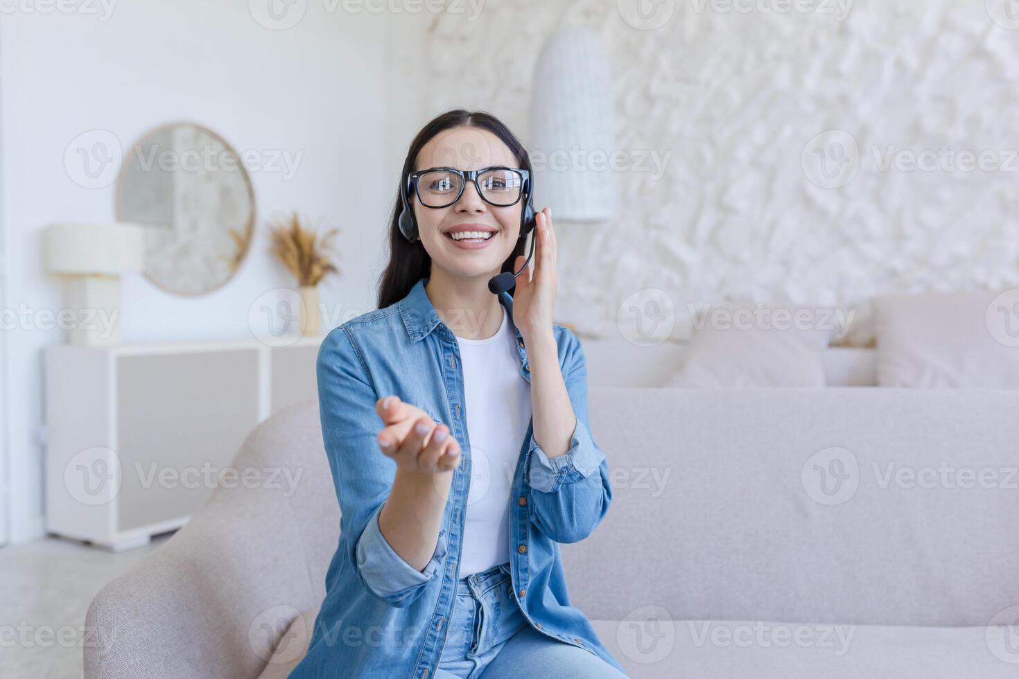 Beautiful young woman in headset sitting at home on sofa in front of camera and vlogging, recording blog, conducting webinar, growth and development trainings, pointing at camera, smiling. photo