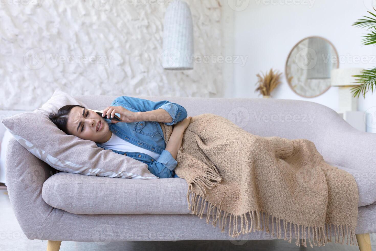 A young woman is lying on the sofa at home, holding her stomach and calling the doctor. Suffers from severe abdominal pain, menstrual pain, pregnancy problems, miscarriage, poisoning. photo