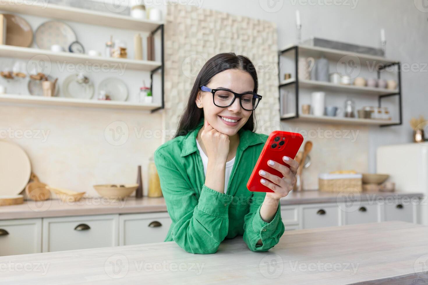 Young woman standing in kitchen at home, leaning on table and using mobile phone, chatting, checking social networks, sending messages, texting with friends, boyfriend. photo