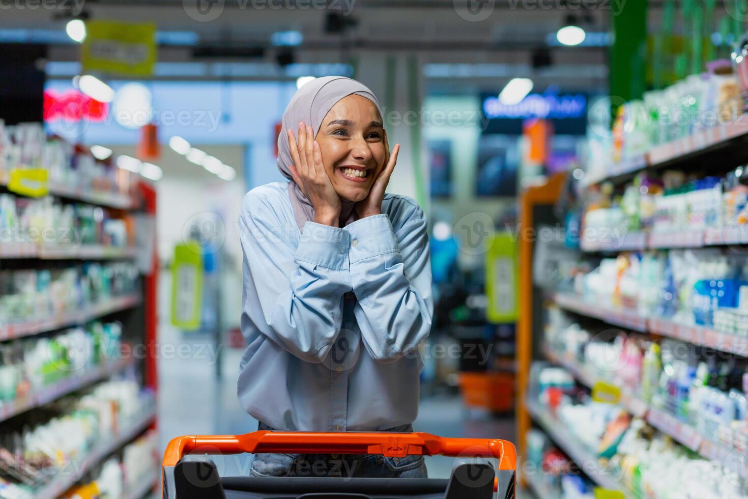 Portrait of successful satisfied woman shopper in supermarket, cheerful muslim woman in hijab with shopping trolley among shelves with goods looking at various shops. photo