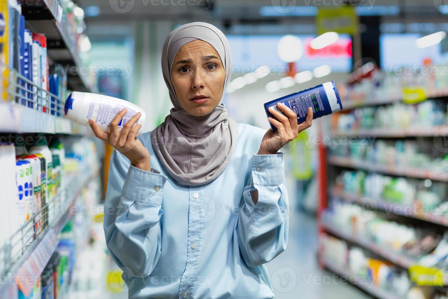 A young Muslim woman in a hijab is standing in a supermarket in the household chemicals department and holding two shampoos. He looks confusedly at the camera, chooses, advertises, shows. photo