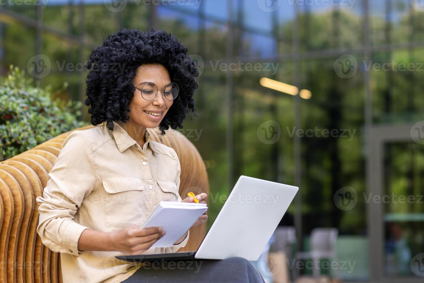 Smiling young African American female student sitting on a bench on a university campus studying online using a laptop. Writes in a notebook and listens to a lecture. photo