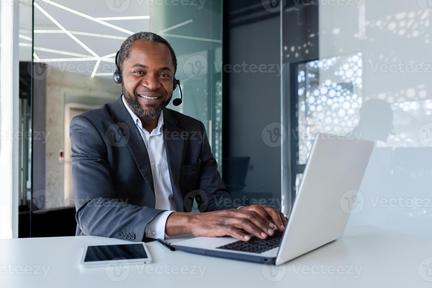 Portrait of african american businessman inside office at workplace, mature man using headset phone and laptop for remote consultation with clients, smiling and looking at camera. photo