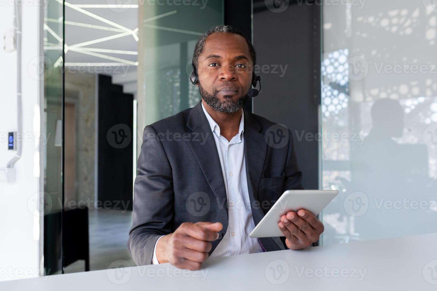 Portrait of a serious senior african man who works in the office in a headset, sits at the table, looks seriously into the camera, uses a tablet. photo