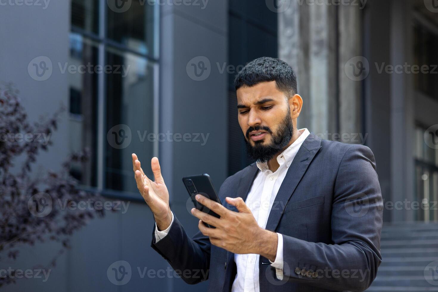 Young arab man businessman read bad news, message. He is standing near the office center, holding the phone and throwing his hands up in displeasure. Close-up photo. photo
