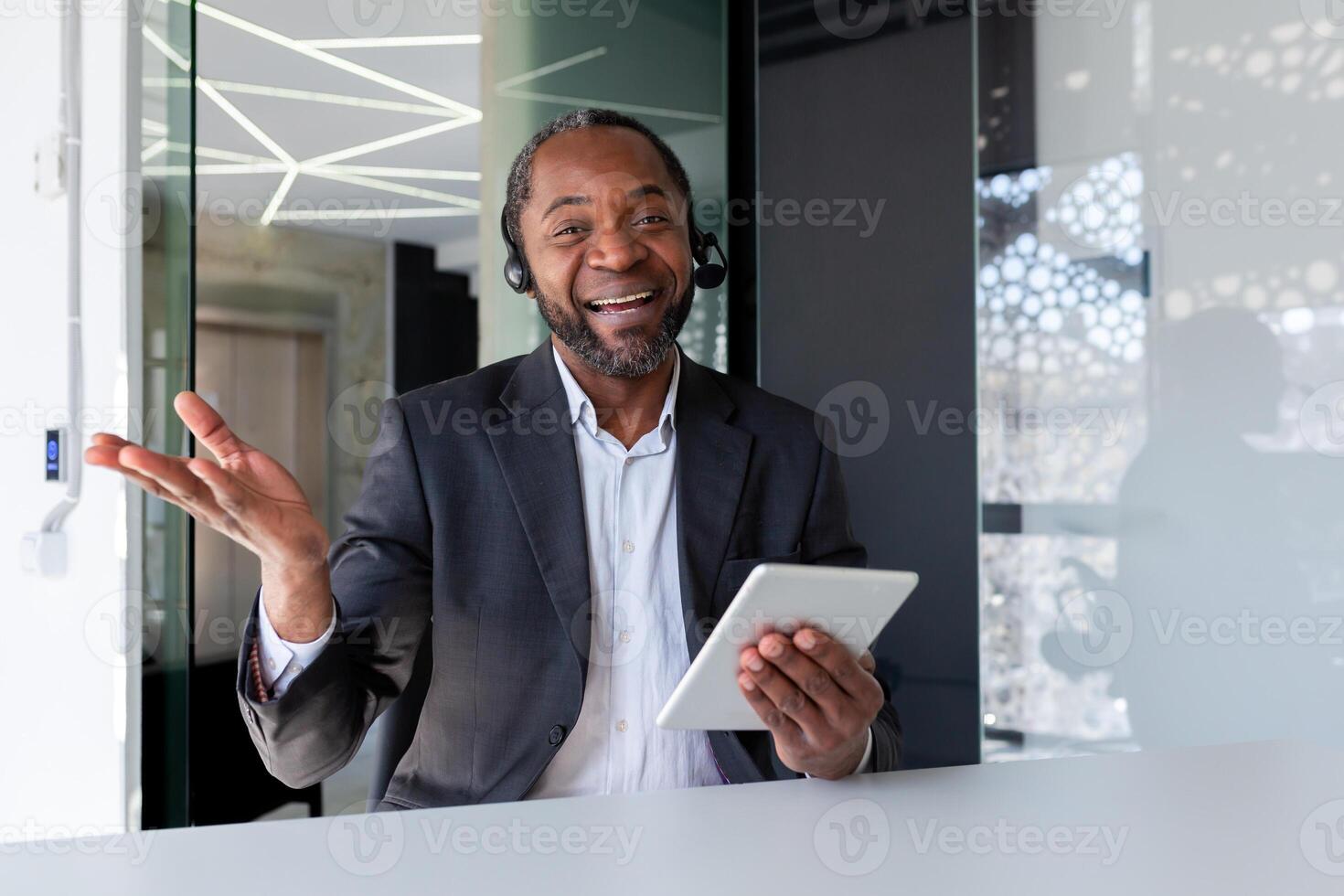 Portrait of a young African-American man working in the office, sitting at a desk in a headset, holding a tablet, smiling and gesturing to the camera. photo