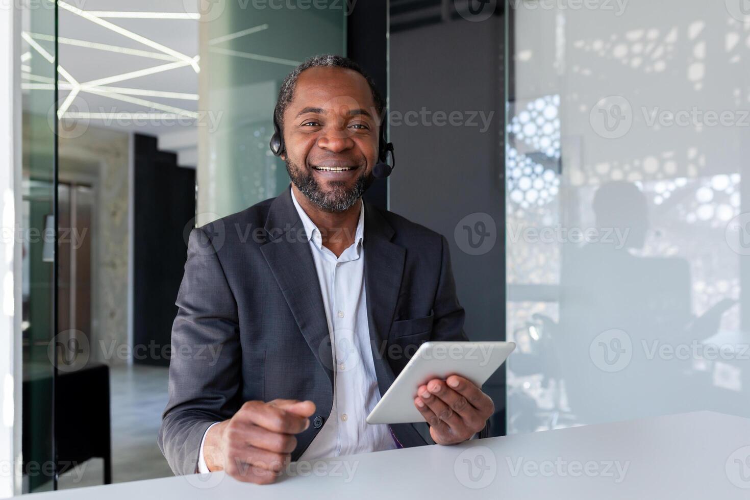 Portrait of support worker inside office at workplace, man smiling and looking at camera, using headset phone and tablet for communication and consulting customer support. photo