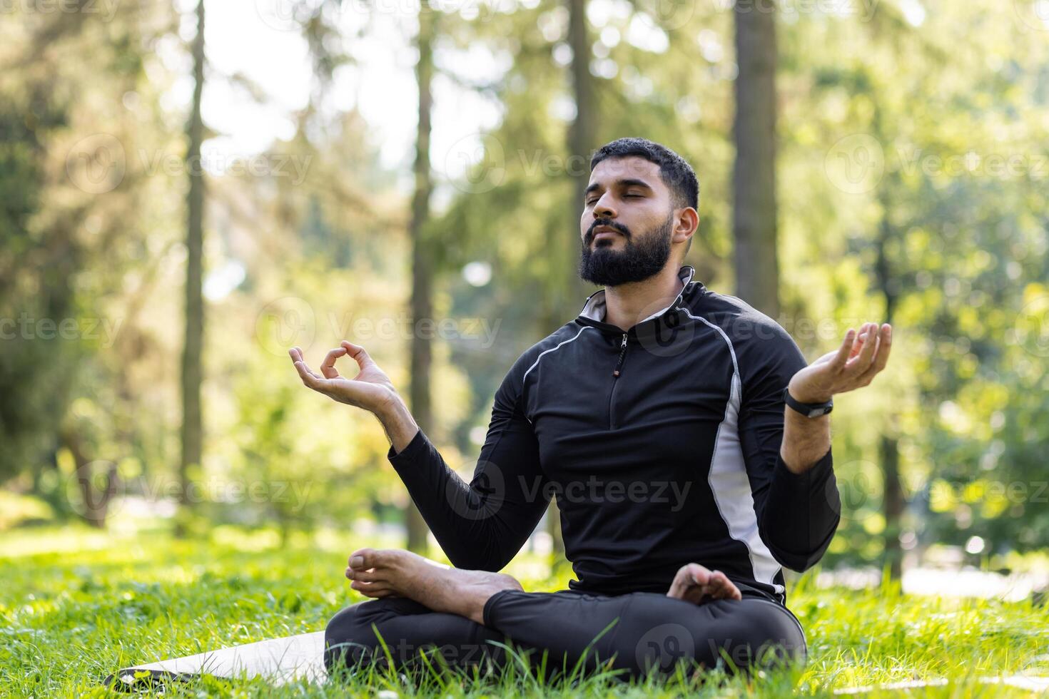 Young Muslim man doing yoga in the park. Sitting on a mat in the lotus position and meditating with closed eyes. photo