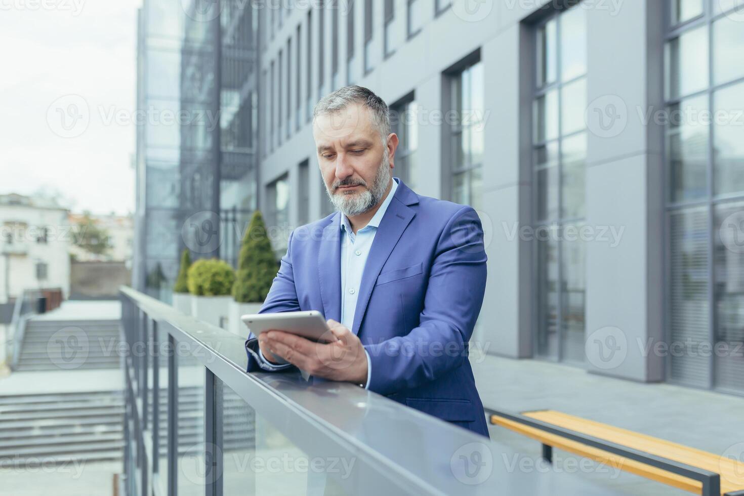 Successful and serious pensive senior gray haired man outside office building using tablet computer, businessman investor reading news from tablet photo