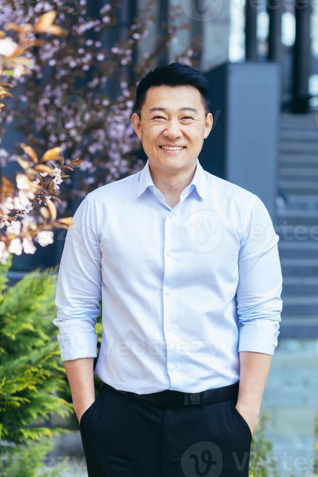 Vertical frame, Asian man in white shirt looks at camera and smiles, hands in pockets photo