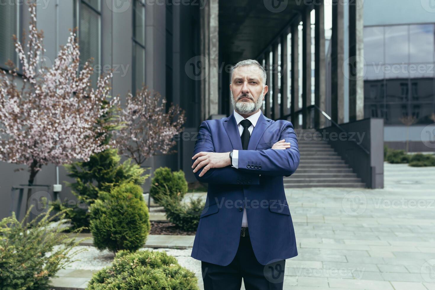 Portrait of successful thinking experienced senior businessman, gray haired male boss in business suit outside office, looking at camera with crossed arms photo