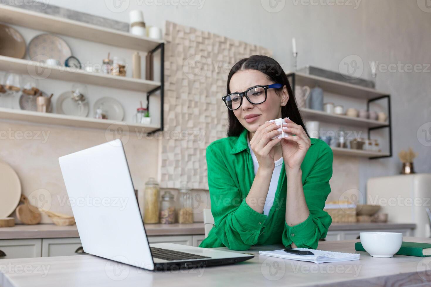 A young woman in glasses and green clothes is sitting at home in the kitchen at the table watching a movie, series, program from a laptop. She is crying, holding a napkin in his hands, wiping his tears. photo