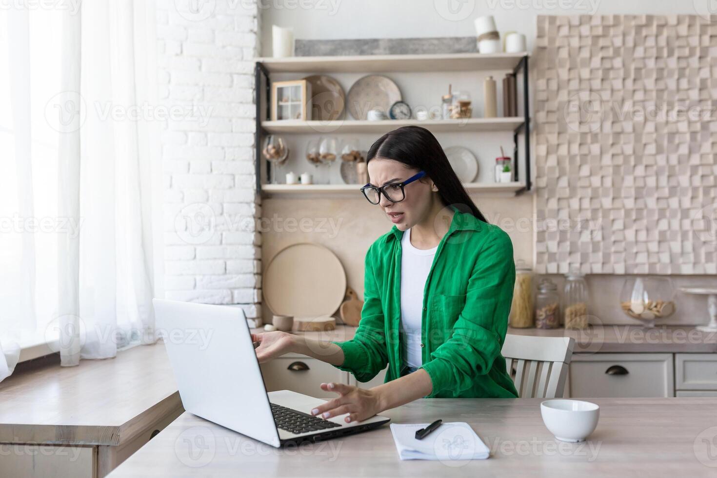 Deadline. A beautiful young business woman in glasses sits at the table in the kitchen, works with a broken laptop, a computer malfunction, waves her hands unhappily, points to the monitor. photo