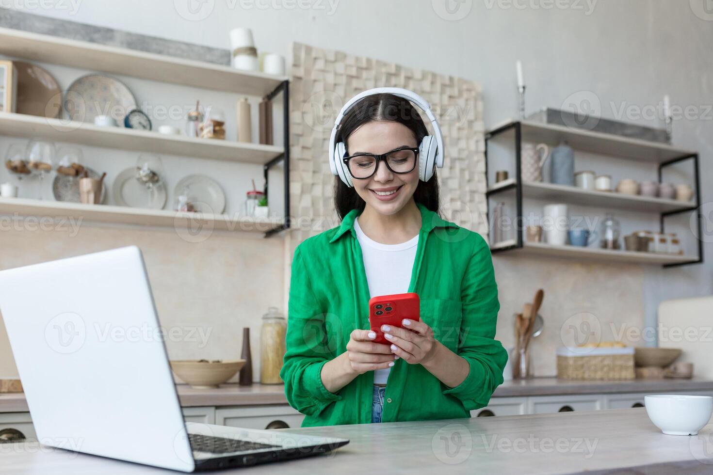 A young beautiful female student is studying in headphones, studying online at home from the phone, listening to a podcast, a lecture. Sitting at a table with a laptop photo