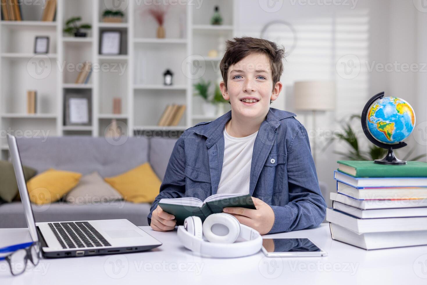 Portrait of a schoolboy at home in the living room, the boy is studying sitting at the desk, reading a book and doing homework, the teenager is smiling and looking at the camera. photo