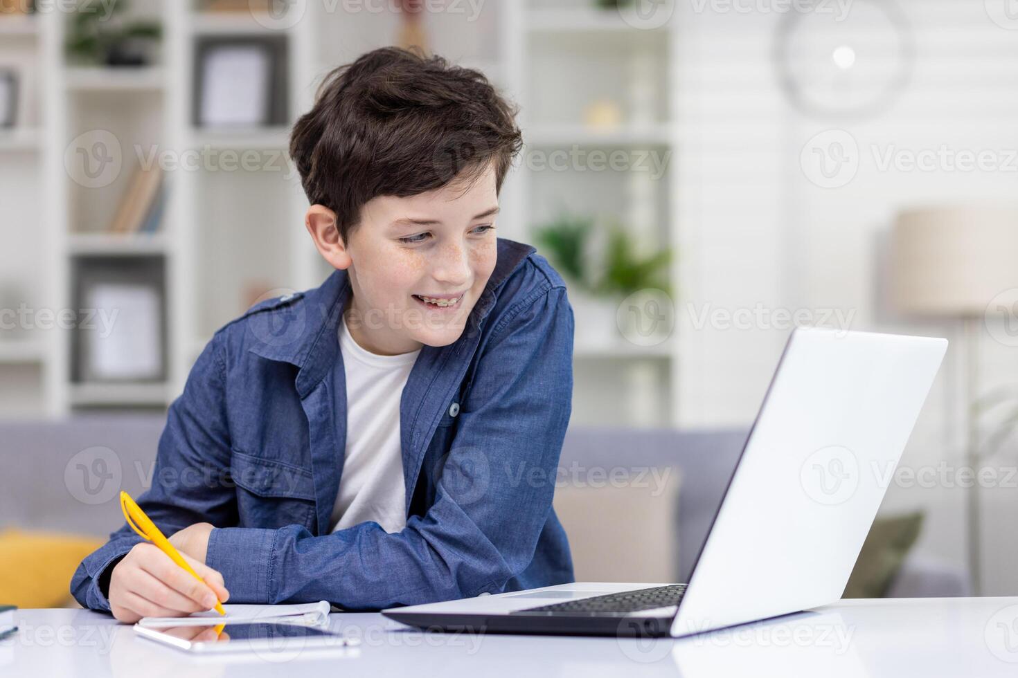 A young guy, a student at home, sits at a desk with a laptop, does homework and studies, smiles contentedly, records online courses, remote learning. photo