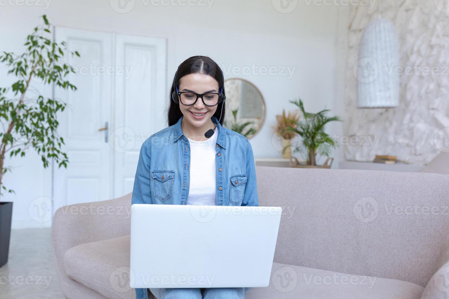 Young beautiful female student studying online at home with laptop on in headphones. He sits on the couch in jeans and glasses, talks via link, listens to lectures, smiles. photo