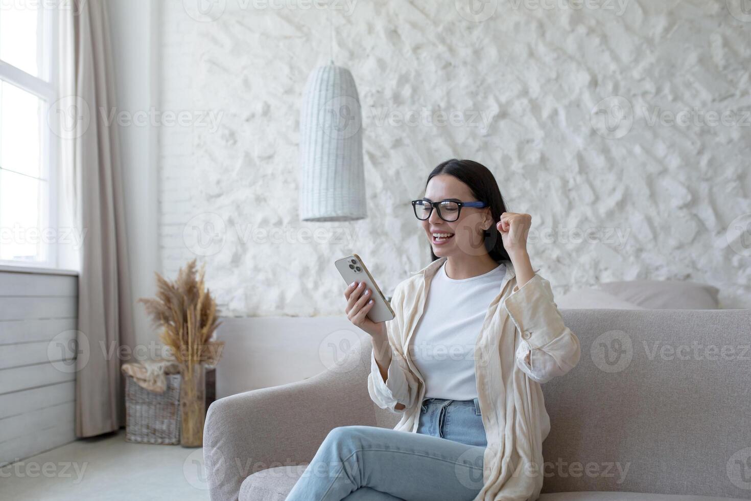 Happy young woman sitting on sofa at home and holding phone. got good news. Got a successful job, passed an interview, received a message. He rejoices, shouts with joy, points with his hand yes. photo