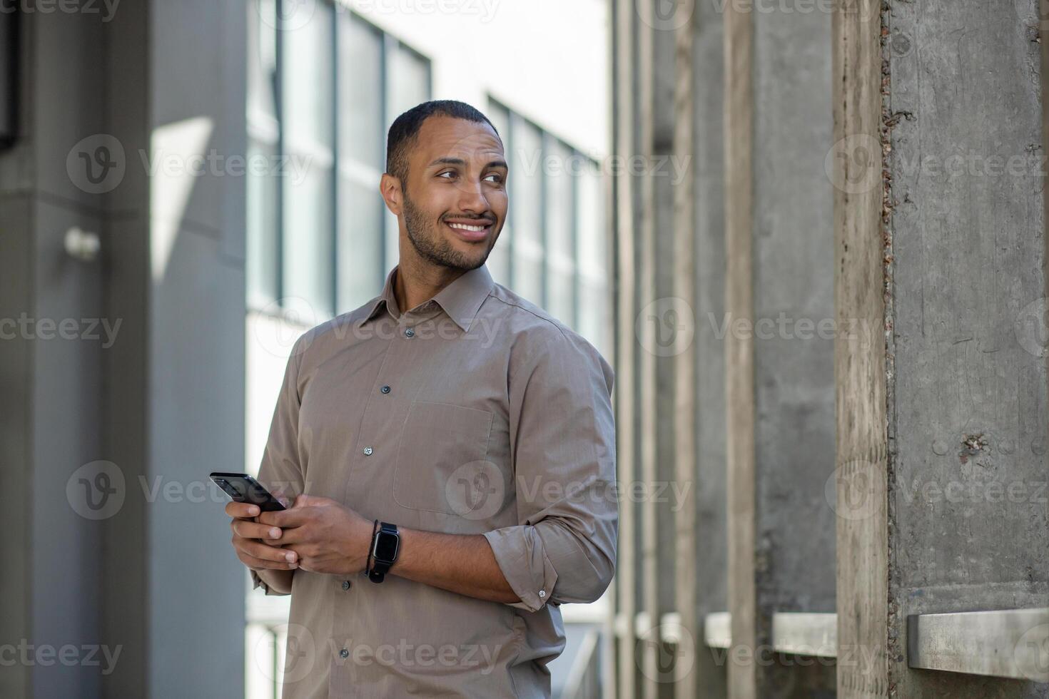 Young joyful successful man looking to the side, satisfied businessman with phone in hands walking around the city outside office building, Indian programmer in shirt. photo