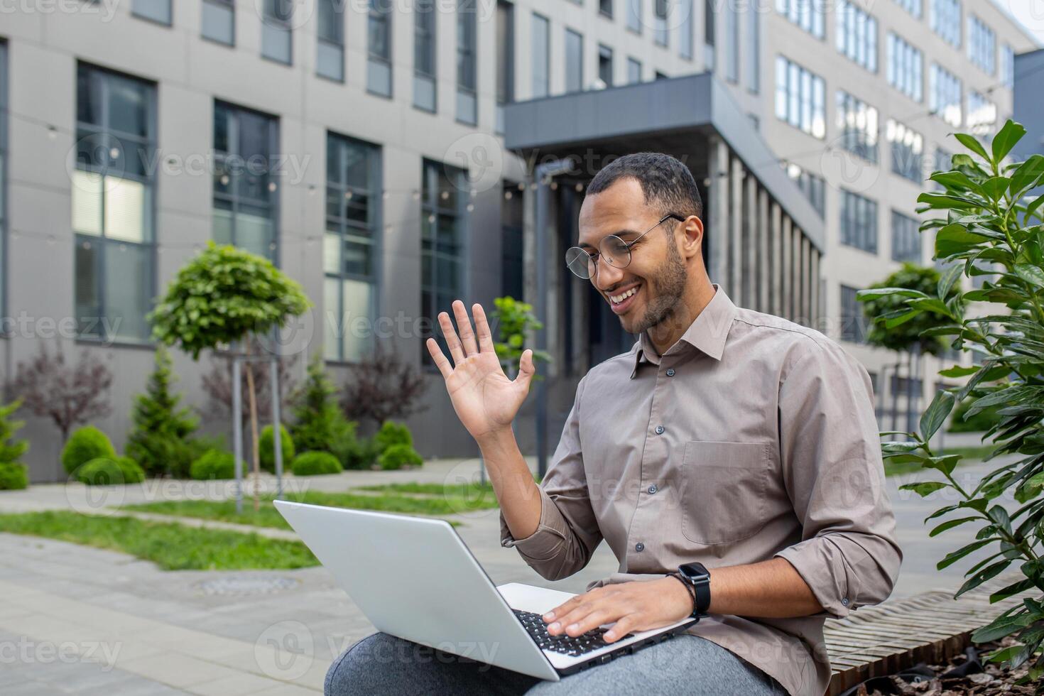 Businessman using laptop for call, man sitting on bench outside office building, waving to interlocutor, greeting gesture. photo