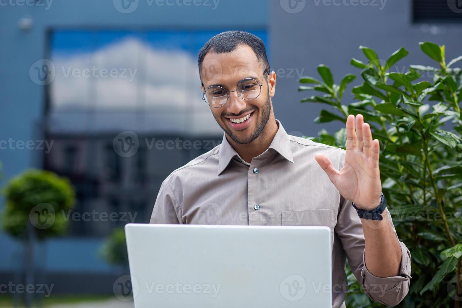 Smiling man in glasses using laptop outdoors, waving at screen, call concept, friendly professional meeting. photo