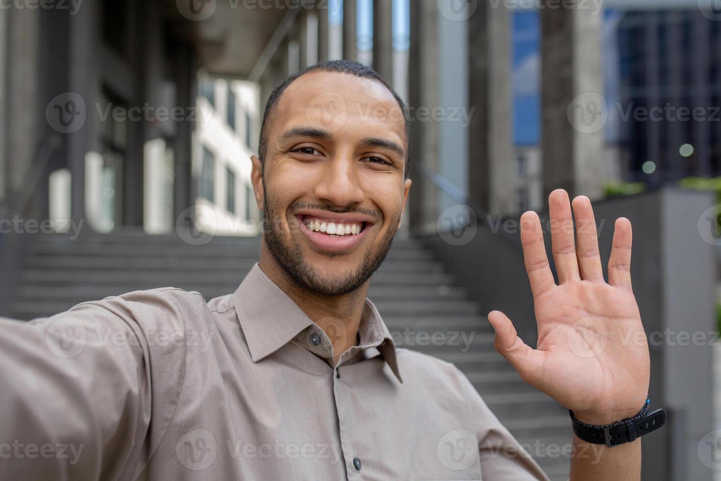 Cheerful young man taking a selfie, waving hand in greeting with a bright smile, urban steps background. Concept of friendliness and technology. photo