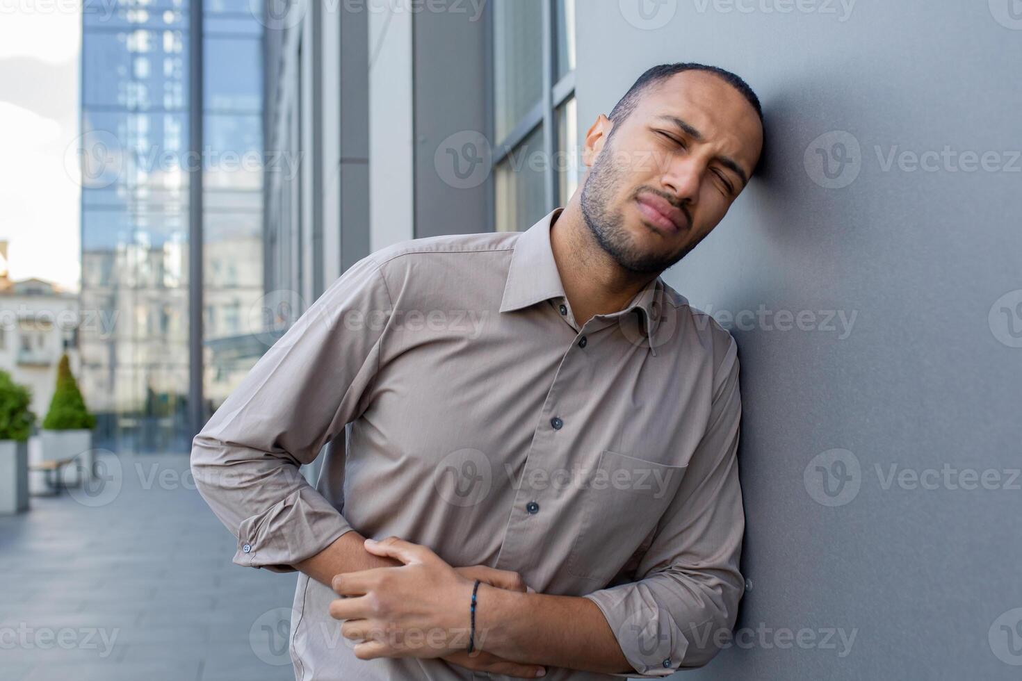 Man in pain outdoors, clutching his side and grimacing, conveying a sense of discomfort or abdominal ache against an urban backdrop. photo