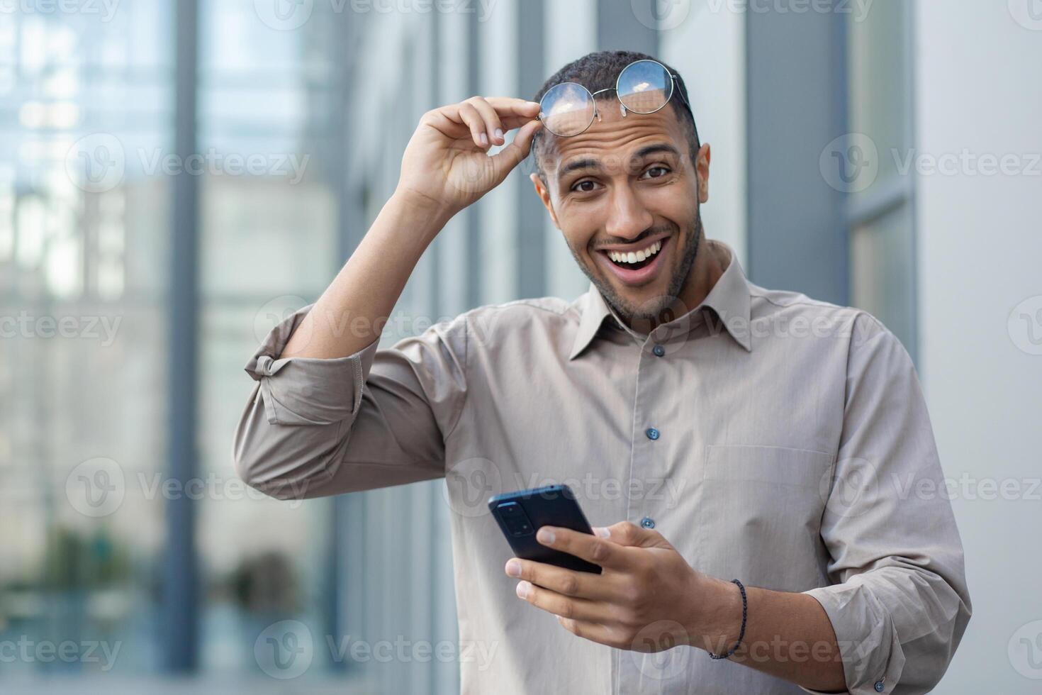 Surprised joyful man looks at the camera, businessman from outside the office building with the phone in his hands walks around the city. photo
