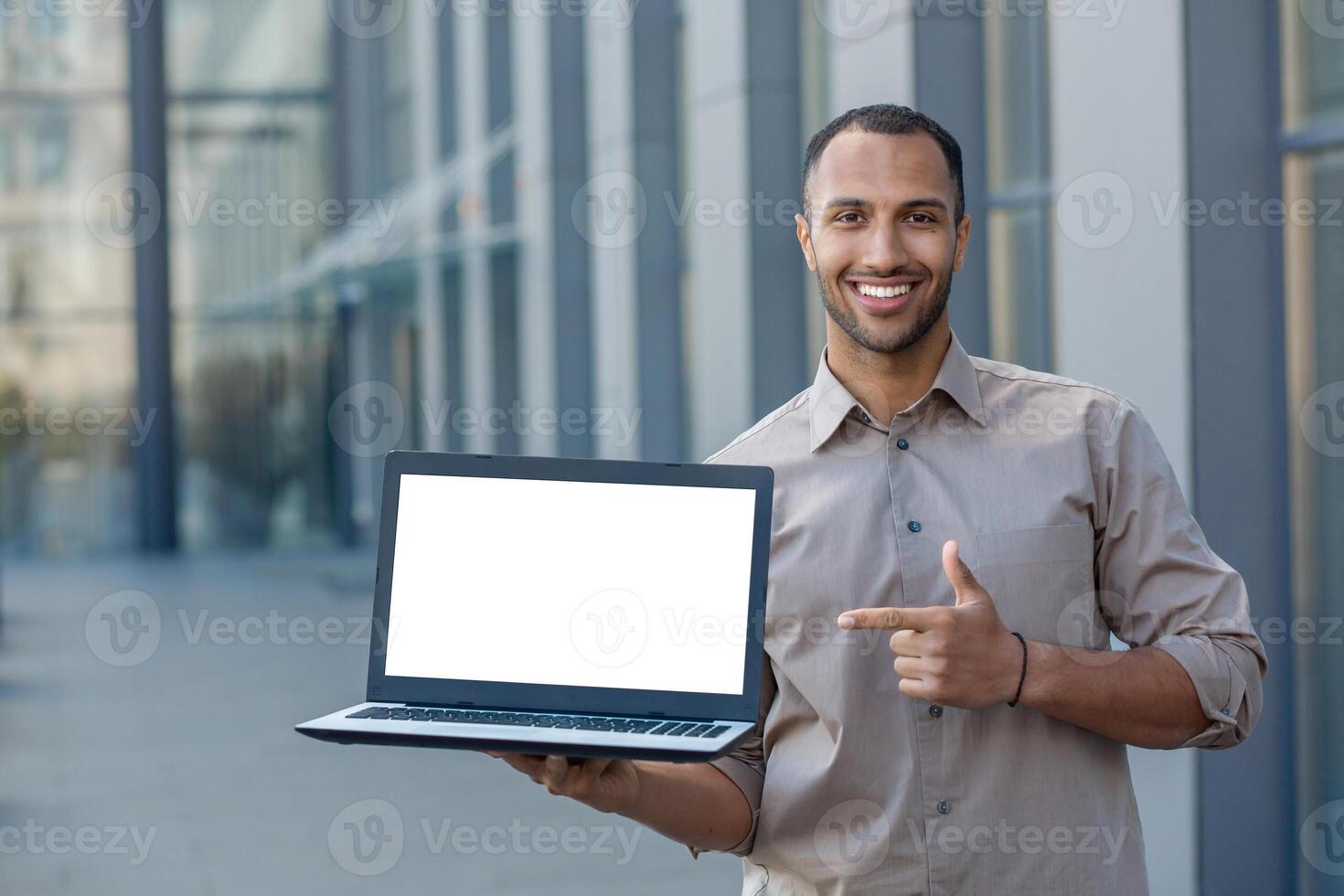 Businessman successful worker outside office building reaching and looking at camera, showing white laptop screen, with space for writing. photo