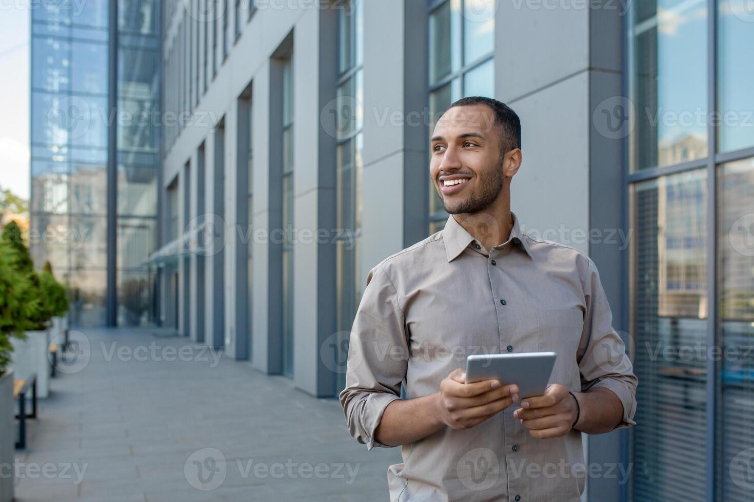 A businessman with a tablet computer in his hands from the outside of an office building, the man is smiling and looking away. An employee in a shirt walks outside an office building, uses an app. photo