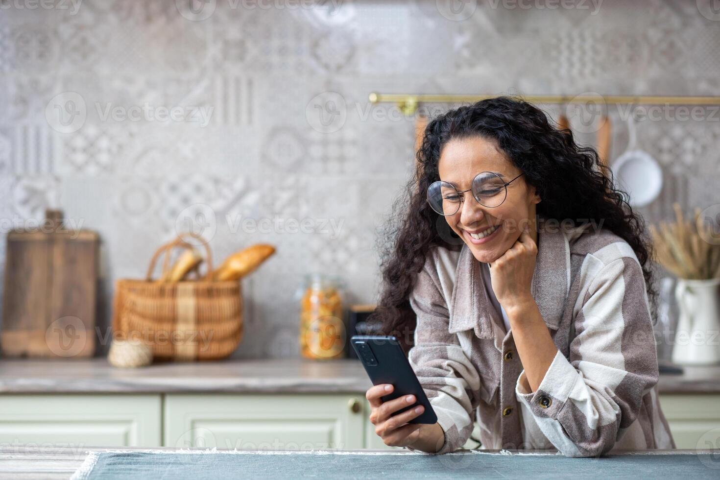 A young beautiful woman at home in kitchen smiles contentedly, browses social networks, online Internet pages, holds phone in r hands. Hispanic with curly hair , using an app on smartphone. photo