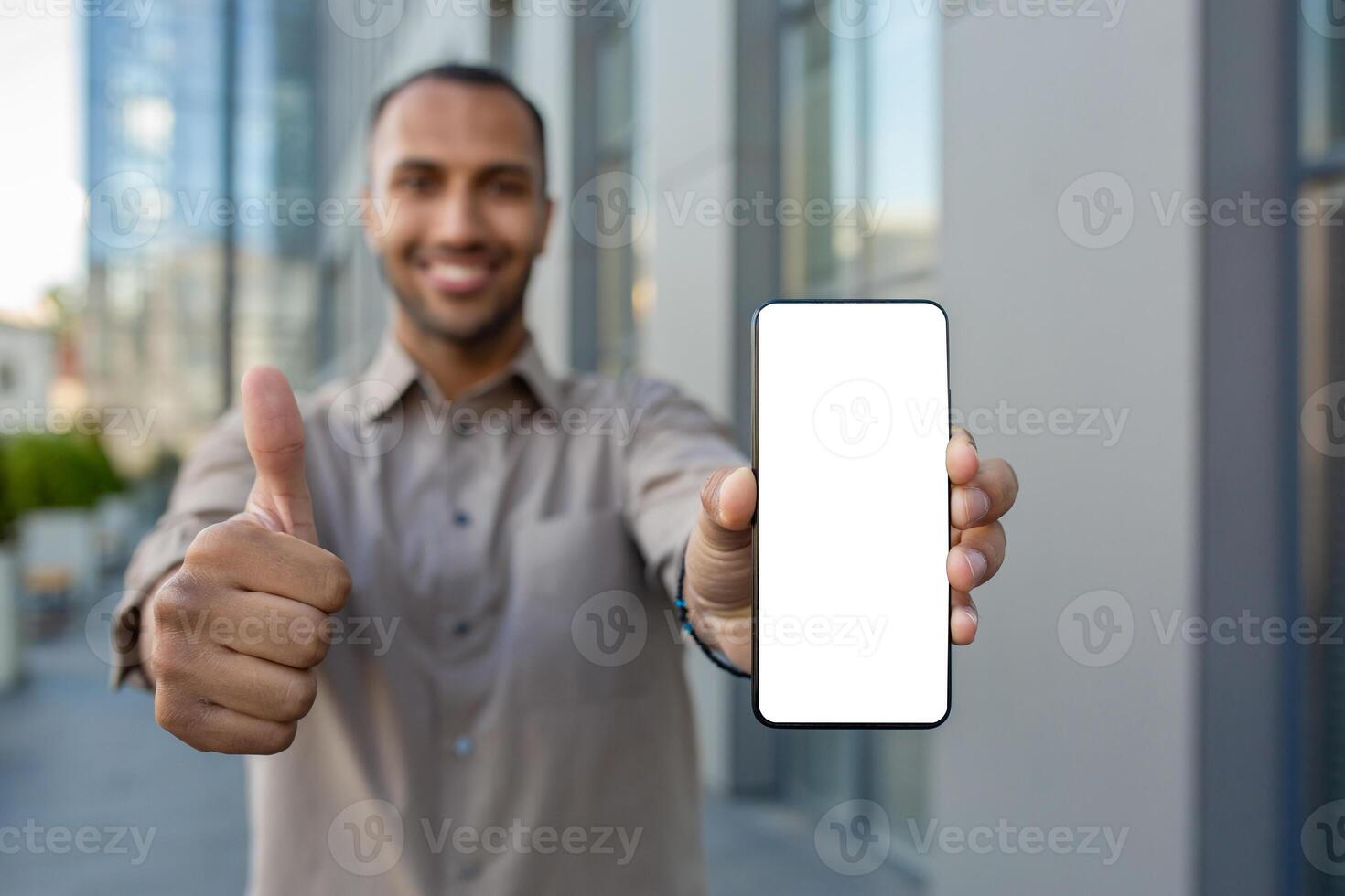 Young successful man showing white phone screen to camera, businessman recommending app, looking happily at camera, outside office building. photo