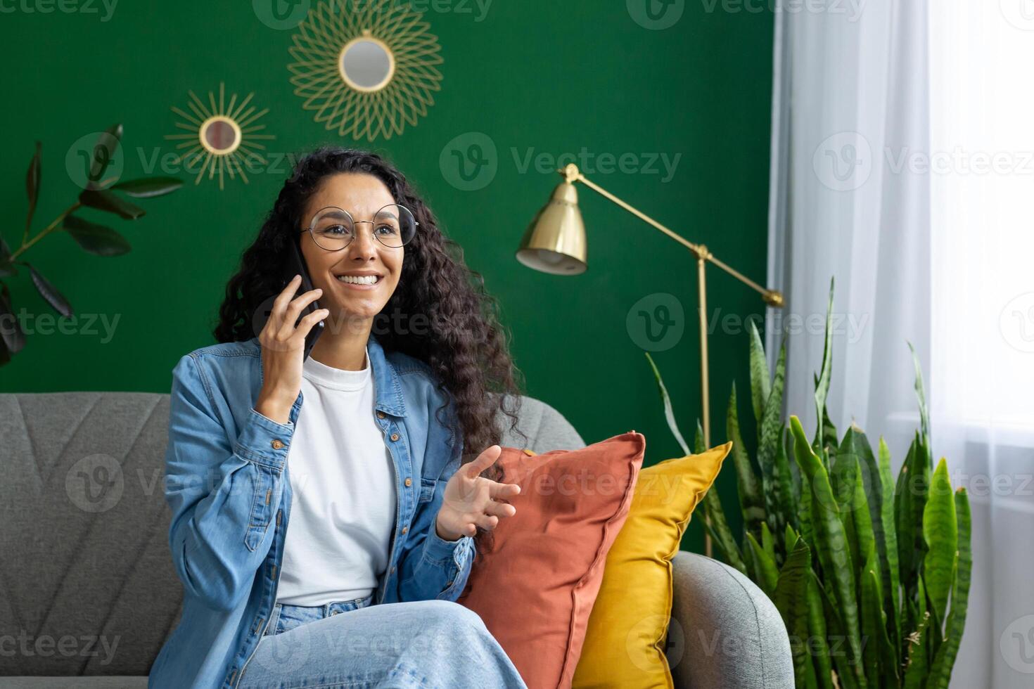 un alegre mujer se involucra en un conversacion en su teléfono inteligente mientras sentado cómodamente en su vibrante vivo habitación, rodeado por decorativo plantas de interior foto