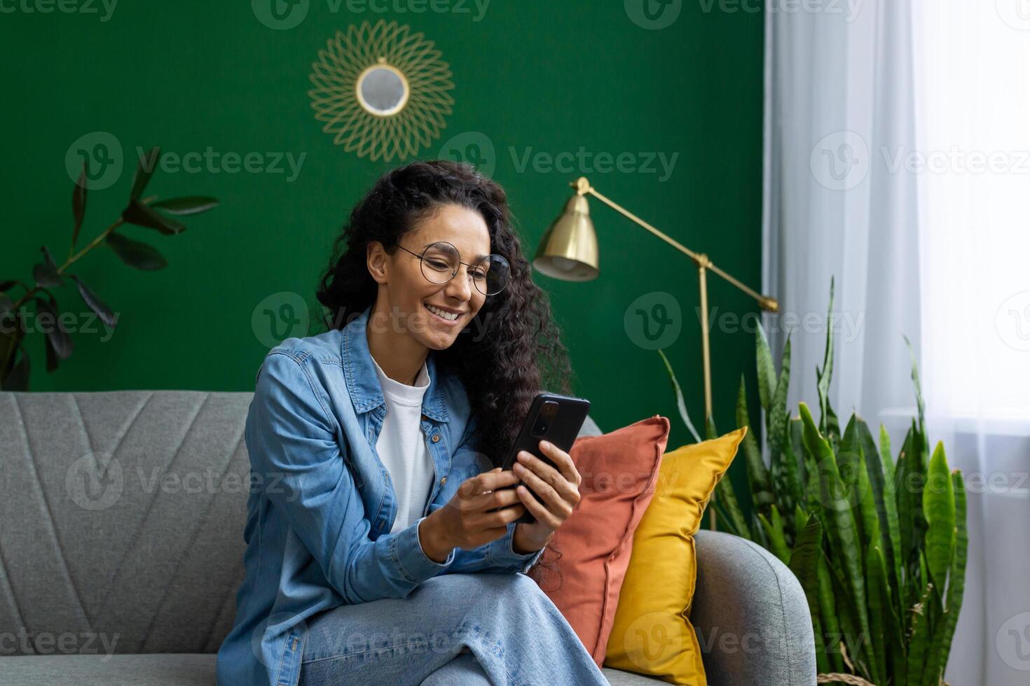 Beautiful Hispanic woman at home on sofa in the living room, the woman smiles contentedly holds the phone in her hands, uses applications on the smartphone, browses the Internet and social networks. photo