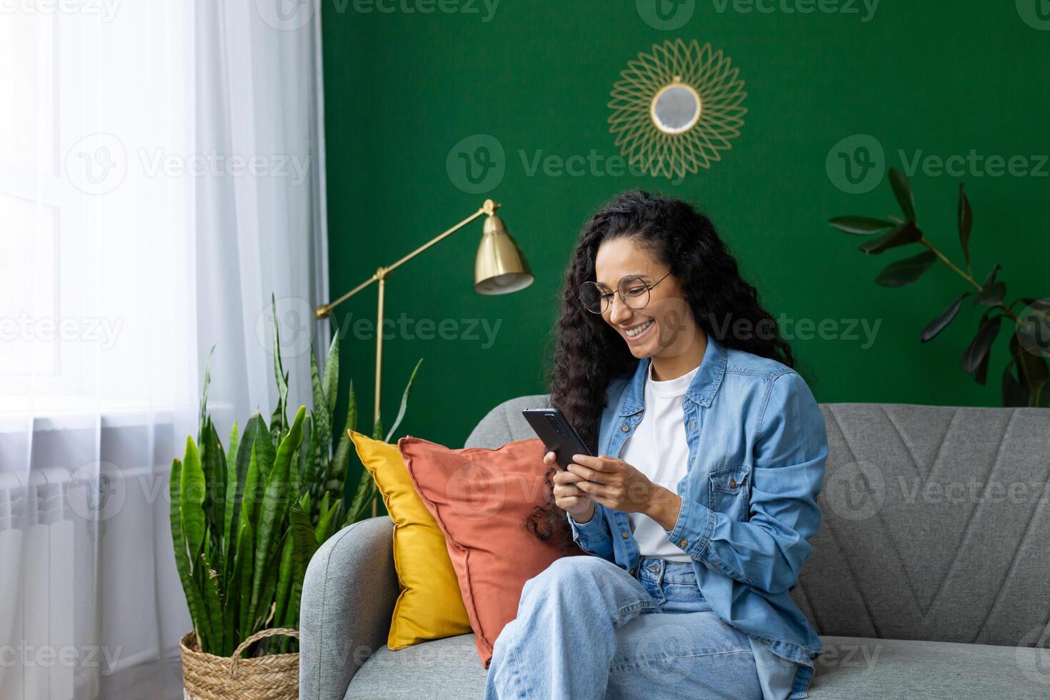 Beautiful Hispanic woman at home on sofa in the living room, the woman smiles contentedly holds the phone in her hands, uses applications on the smartphone, browses the Internet and social networks. photo
