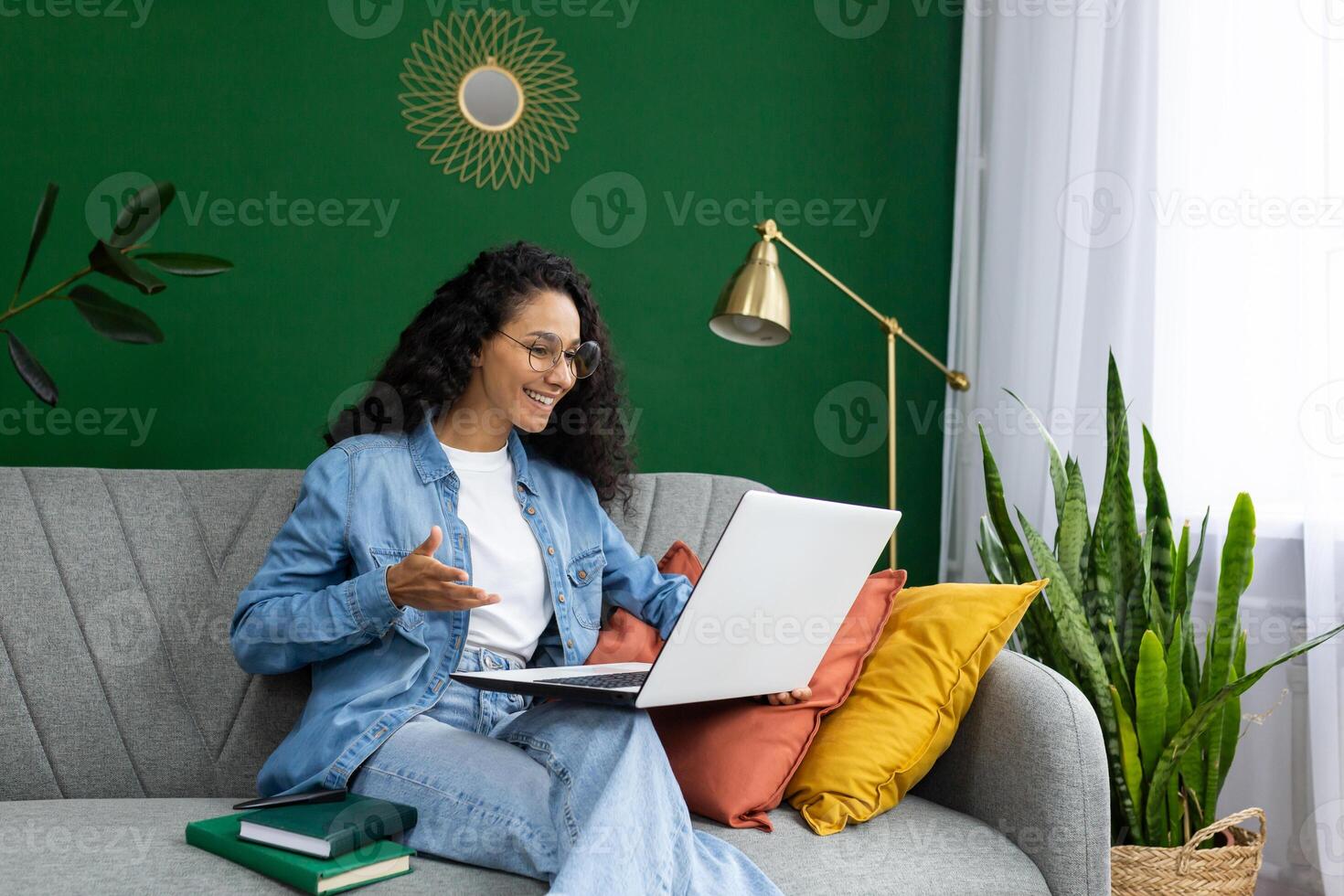 sonriente mujer en lentes teniendo un llamada en ordenador portátil a hogar, expresando alegría y comodidad en un acogedor vivo habitación ajuste. foto