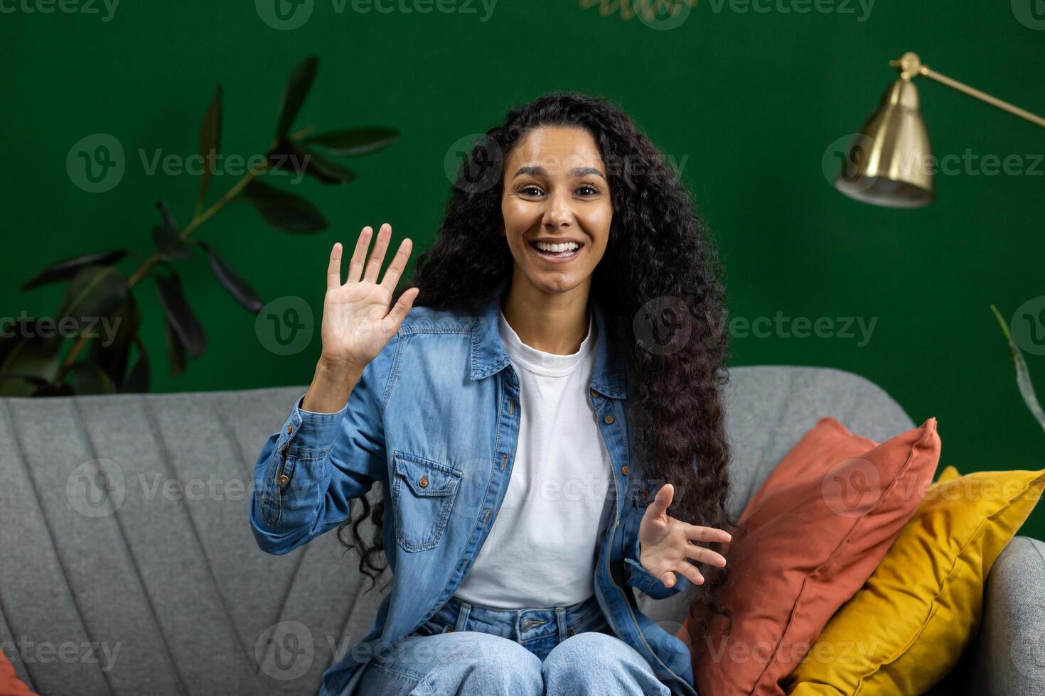 Young beautiful woman at home on sofa looking at web camera, talking with friends remotely, smiling with satisfaction and gesturing with hands, using call on computer, waving hand greeting. photo