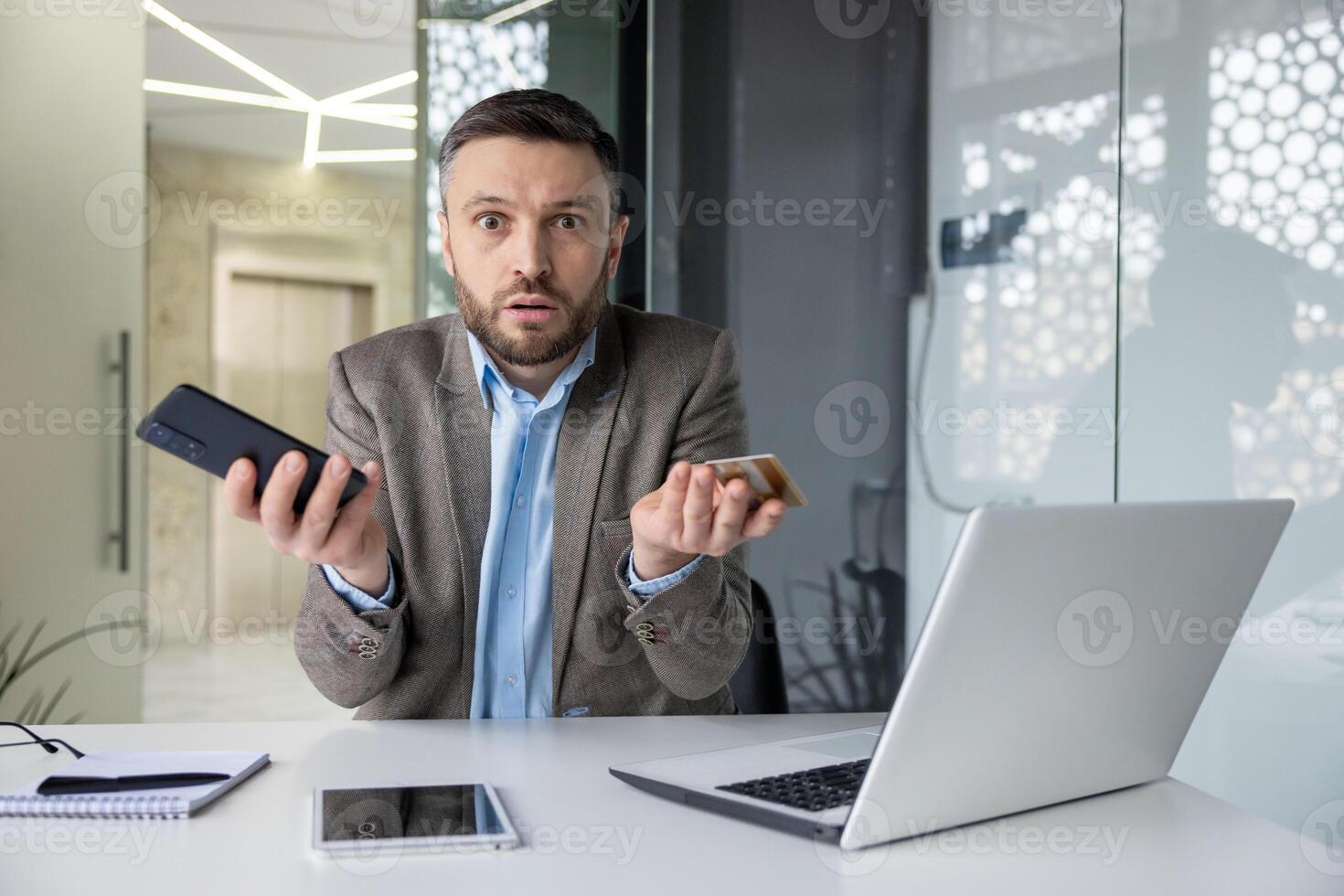 A businessman in a modern office appears confused while holding a smartphone and multiple credit cards. He's dressed in a professional suit in a well-lit setting. photo