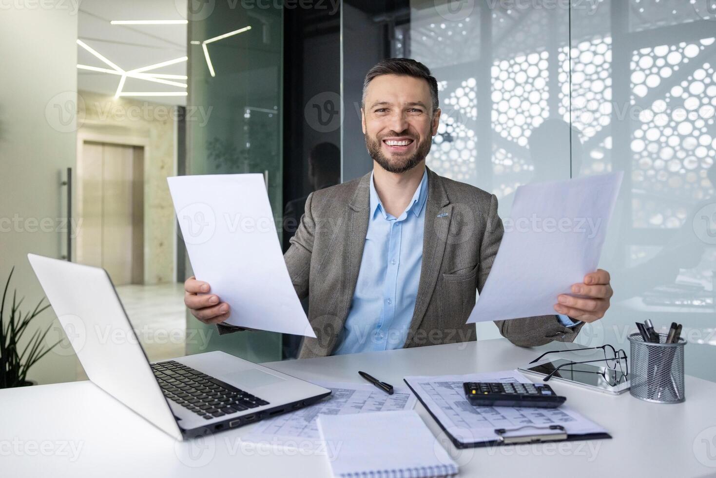 Smiling businessman sitting at a desk in a modern office, holding papers, with a laptop and calculator nearby. Portrays a productive and positive work environment. photo