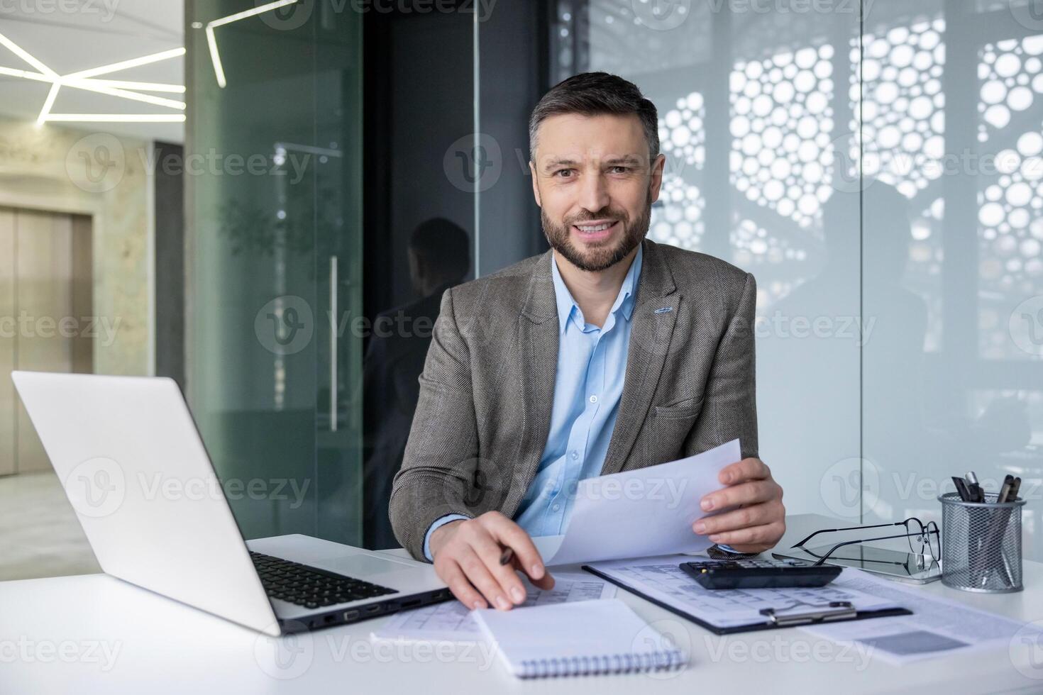 Confident middle-aged businessman in a stylish modern office working at his desk. He reviews documents while engaging with a laptop, portraying professionalism and focus. photo