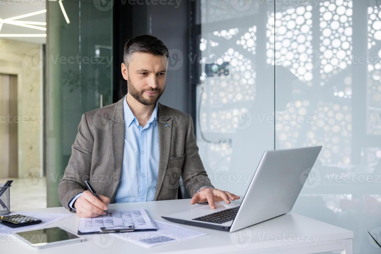 A focused business man reviewing documents while working on his laptop in a modern office setting, with natural light backlighting the scene. photo