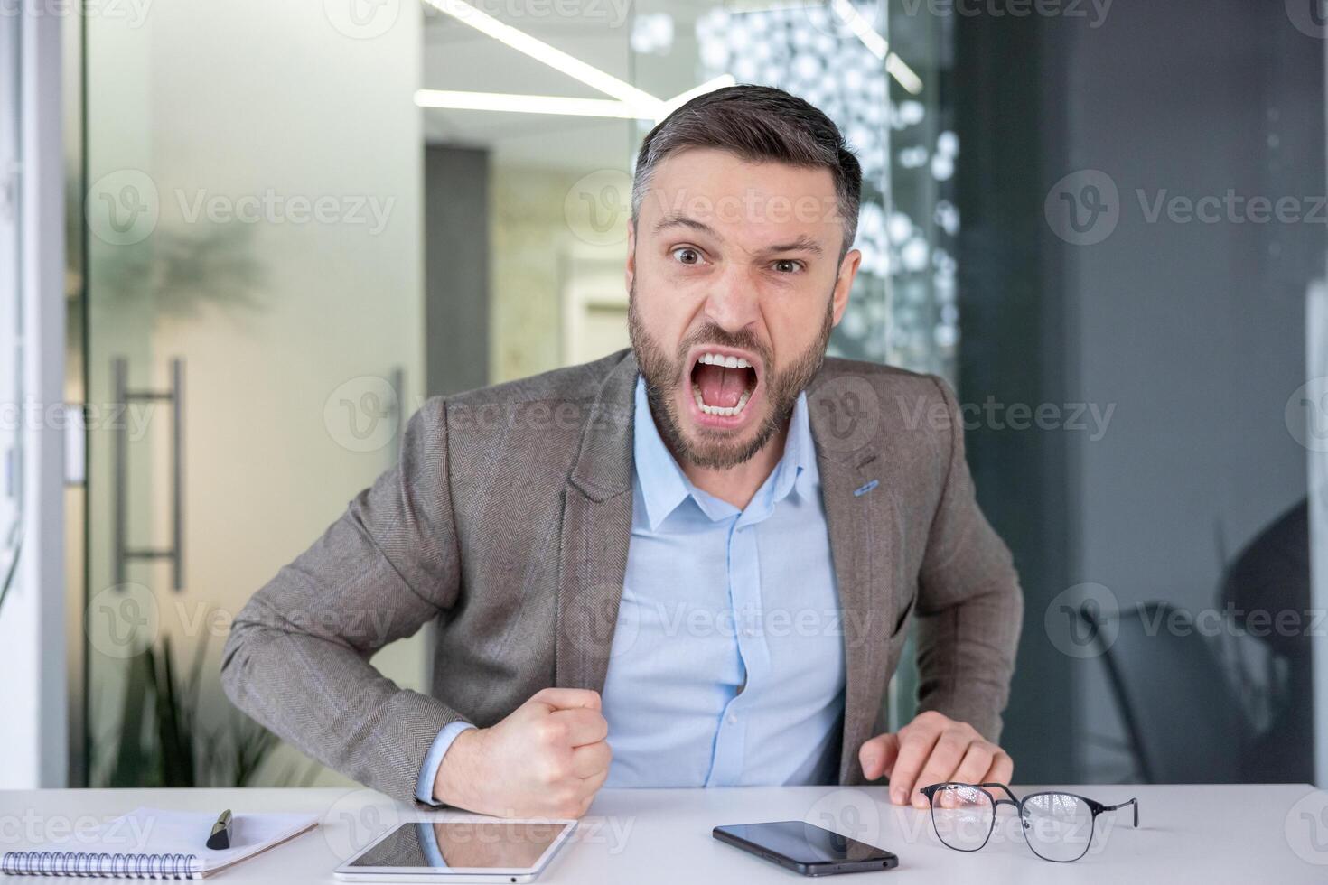 An angry male office worker screaming with clenched fists, showing strong emotion and workplace stress against a corporate interior background. photo