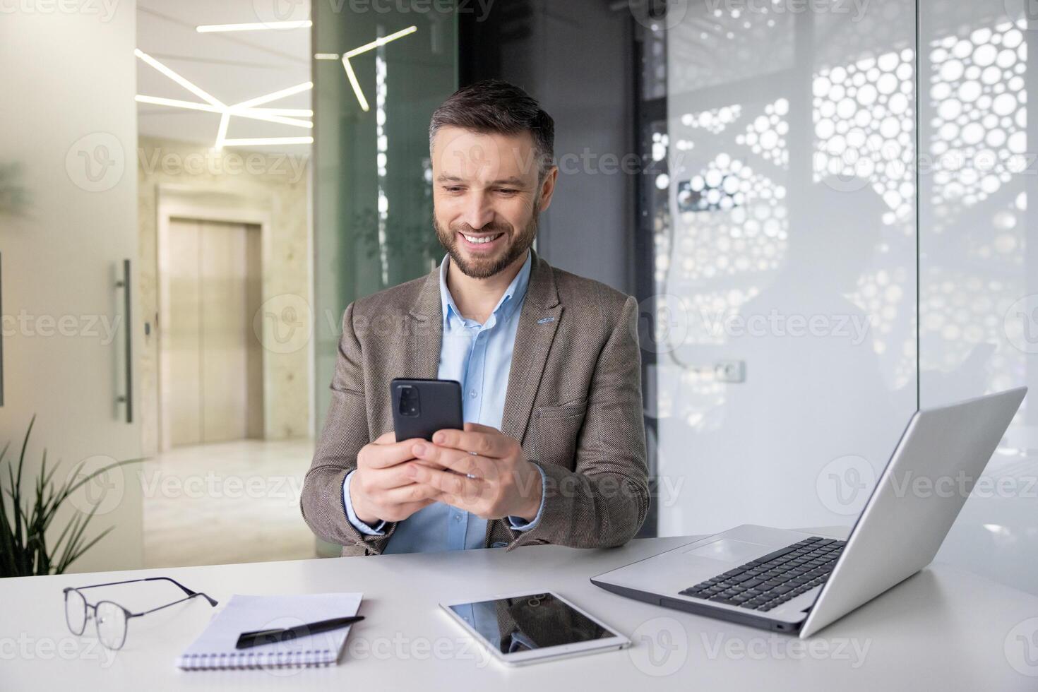 Joyful smiling satisfied businessman typing text message on phone, man in business suit browsing social media and using app on smartphone, sitting inside office with laptop at workplace. photo