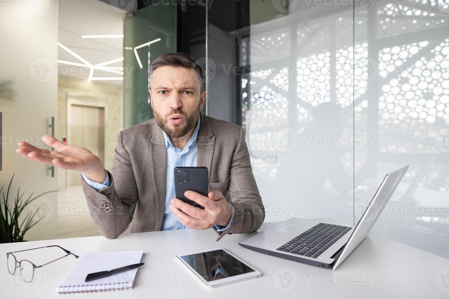 Confused businessman with phone at desk expressing frustration, surrounded by tech gadgets in a modern office setting. photo