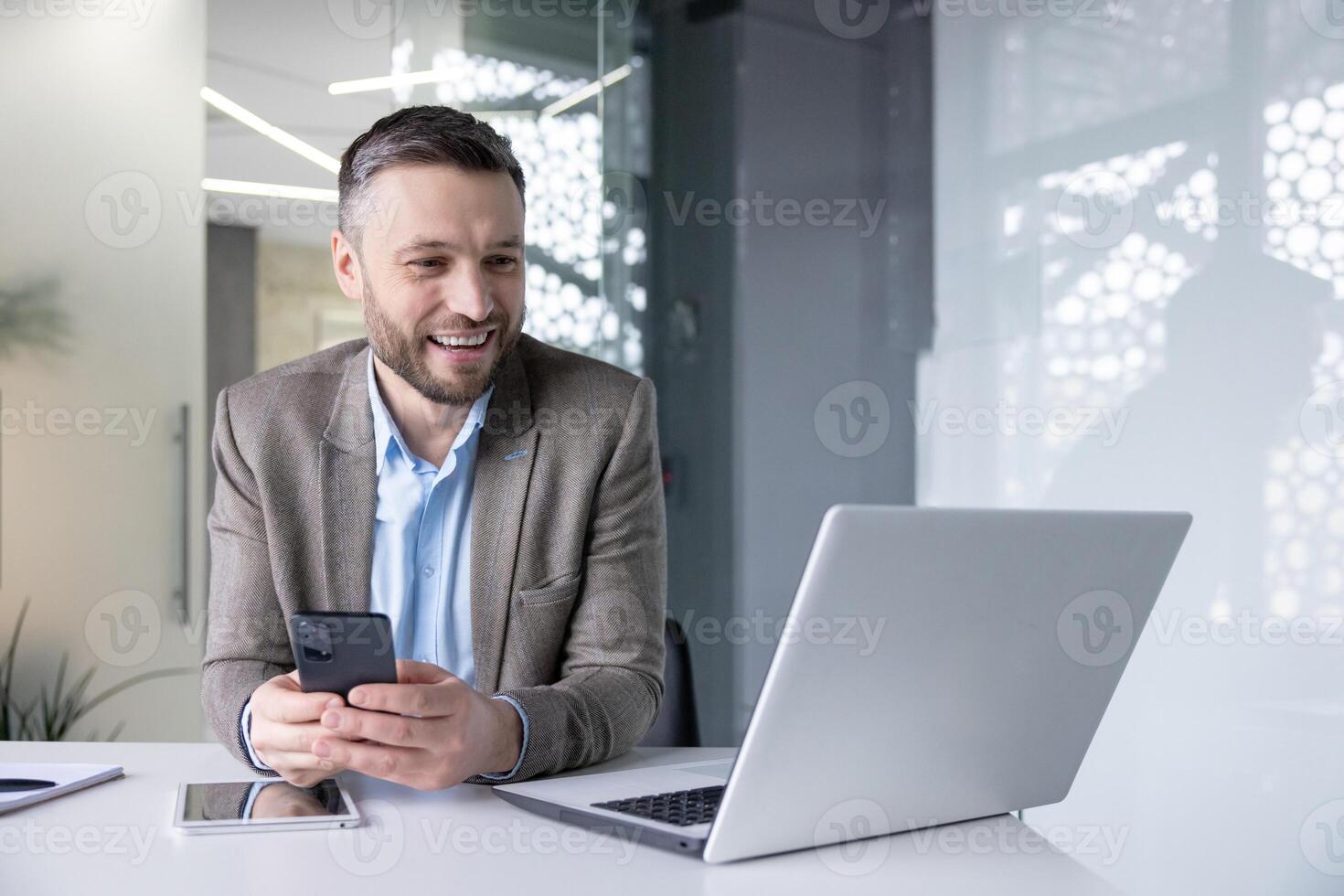A young male businessman sits at a desk in the office, holds a phone in his hands and communicates via call on a laptop. photo